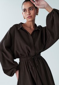 Rjava obleka s srajčnim ovratnikom, dolgimi rokavi in zategnjenim pasom. Ima razkošne rokave in mehak, teksturiran material.