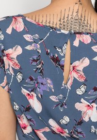 Květinová halenka s modrým pozadím, zdobená růžovými a fialovými květy, s krátkými rukávy a zadním zapínáním na knoflík ve tvaru průduchu.