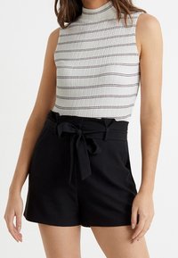 Top bianco senza maniche con strisce grigie orizzontali; abbinato a pantaloni corti neri a vita alta con cintura legata e texture liscia.