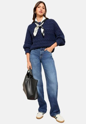 Maglione navy a lavorazione cable-knit con maniche a sbuffo, abbinato a jeans a gamba larga blu chiaro e una borsa a tracolla in pelle nera. Sneakers bianche con strisce verdi.