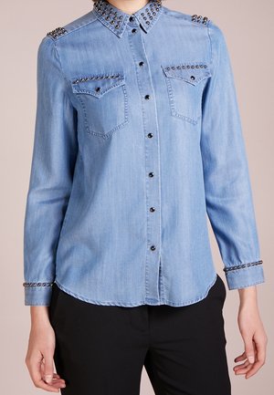 Chemise en denim bleu clair à manches longues avec boutons noirs et clous métalliques sur le col, les épaules, les poches poitrine et les poignets, portée avec un pantalon noir.