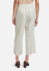 Lichtcrème, wijde broek van zachte stof met een elastische tailleband en twee achterzakken. G gedragen met zwarte haksandalen.