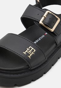 Svarte sandaler laget av syntetisk materiale. Har to brede stropper, en gullspenne og et logoaksent. Tykk, teksturert såle for bedre grep.