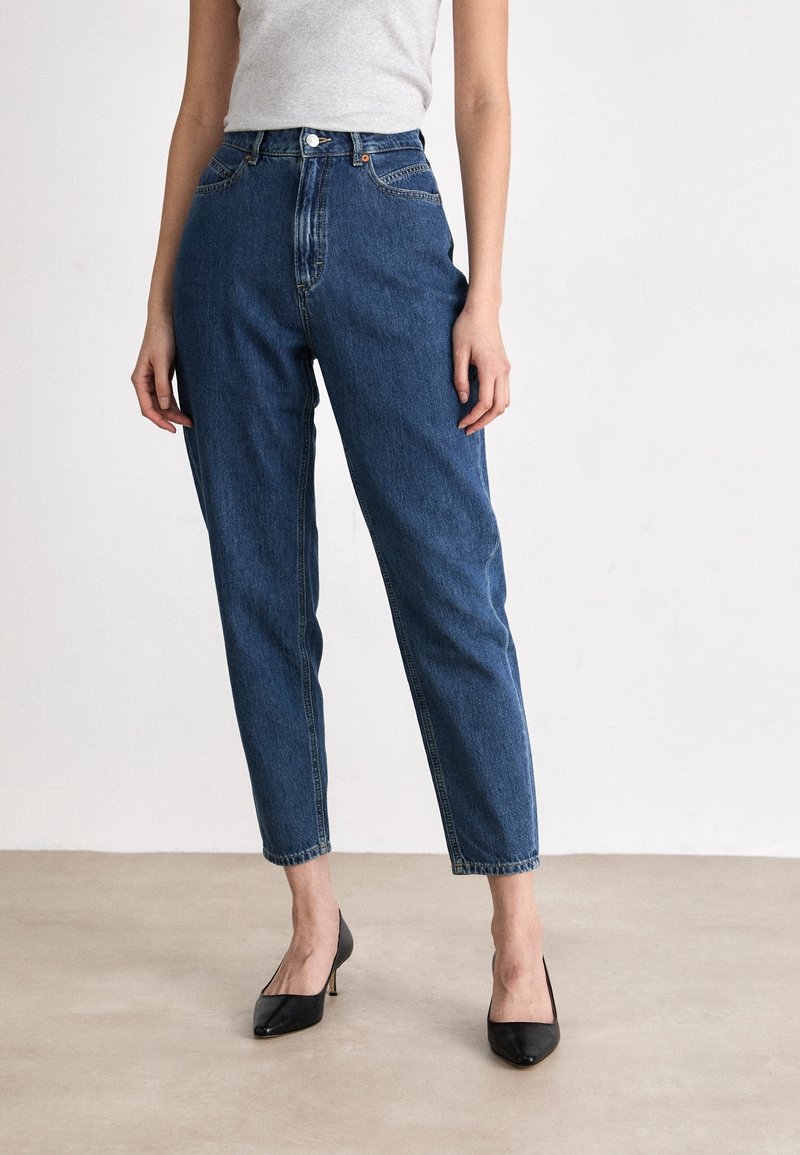 Femme portant un jean bleu taille haute, un haut blanc sans manches et des talons noirs pointus debout sur un sol clair contre un mur blanc uni.