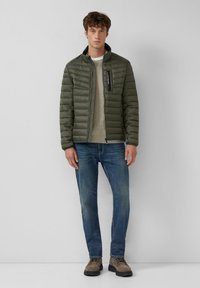 Olivgrüne Daunenjacke mit Reißverschlusstasche, kombiniert mit einem beigefarbenen Pullover, blauen Jeans und grauen Stiefeln, stehend vor neutralem Hintergrund.