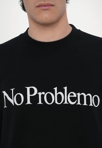 Svart hettegenser med hvitt tekst "No Problemo" i en fet, serif-skrift. Stoffet ser mykt ut med en klassisk ribbet halslinning.