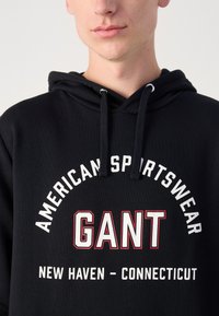 Čierna mikina s kapucňou a šnúrkami, s výraznou bielou a červenou grafikou, na ktorej je nápis "GANT" a "AMERICAN SPORTSWEAR NEW HAVEN - CONNECTICUT."