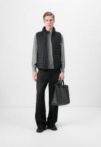 Zwarte donsvest over een grijze trui, gecombineerd met donkere jeans en zwarte schoenen. Houdt een gestructureerde zwarte tote-bag vast met meerdere vakken.
