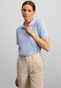 Lys blå polo-shirt med klassisk krave og korte ærmer, parret med beige bukser med en subtil tekstur og knapdetaljer.