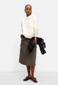 Kvinde står iført cremefarvet sweatshirt, olivengrøn cargo-nederdel, bordeaux flade sko, holder sort jakke i højre hånd mod en ensfarvet hvid baggrund.