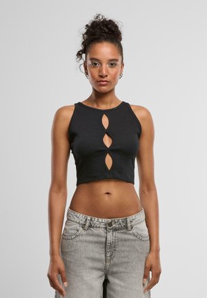 Młoda kobieta z kręconymi włosami w wysokim kokie, ubrana w czarny crop top z wycięciami z przodu oraz jasnoszare, wysokie dżinsy, stoi na tle białym.