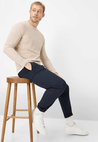 Maglione beige a maniche lunghe, pantaloni navy a vestibilità comoda, sneakers bianche, seduto su uno sgabello di legno. Tessuto liscio, materiale leggero.