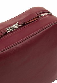 Borsa in pelle bordeaux con una superficie liscia, dotata di una cerniera argentata e un tirante in pelle. Forma rettangolare compatta con bordi cuciti.