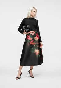 Crna bluza s dugim rukavima u kombinaciji s satenskom suknjom s cvjetnim uzorkom u ružičastim i zelenim nijansama. Model drži crnu clutch torbicu s teksturom.