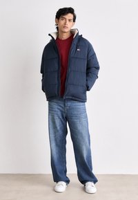 Chaqueta acolchada azul marino con cuello alto y capucha, combinada con un suéter burdeos y jeans azul claro. Se lleva con zapatillas blancas.