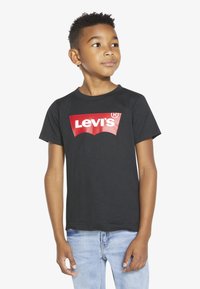 Melnas kokvilnas bērnu T-krekls ar sarkanu Levi's logotipu izliekta dizainā, īsām piedurknēm un klasisku apaļu kaklu. Redzami džinsa džinsi.