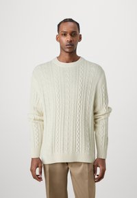 Cream knitted sweater featuring cable patterns, ribbed crew neck, and long sleeves, paired with light brown trousers.
