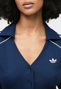 Tumši zils ribots polo krekls ar V-veida apkakli, trīs zilām pogām un baltu apmali apkaklē, kā arī baltu logotipu uz krūtīm.