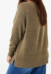 Olivgrüner Pullover mit texturiertem Finish, langen Ärmeln, seitlichen Schlitzen und einem gerippten Saum. Kombiniert mit blauen Denim-Jeans.