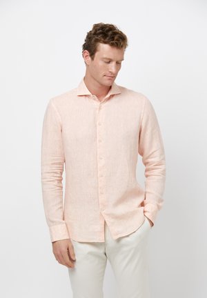 Camisa de manga larga de color rosa claro con una tela texturizada, cierre de botones en la parte delantera y cuello puntiagudo. Llevada de manera casual con las manos en los bolsillos.