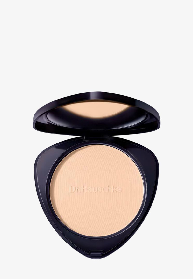Fond de teint compact dans un boîtier triangulaire noir, ouvert pour dévoiler une poudre beige avec l'inscription "Dr. Hauschka" en relief sur la surface.