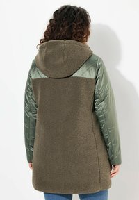 Olivgrön isolerad jacka med en mjuk, texturerad fleecekropp, släta vadderade ärmar och en huva. Inga framträdande mönster eller detaljer.