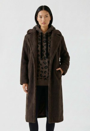 Femme portant un long manteau duveteux marron foncé par-dessus un sweat à capuche marron imprimé léopard avec des cordons noirs, debout avec une main dans la poche.