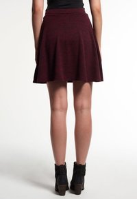 Femme portant une jupe évasée bordeaux et des bottines noires, se tenant de dos avec les jambes visibles contre un fond blanc.