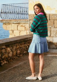 Green striped sweater with pink collar, light blue denim flared skirt, and white lace-up flat shoes. Water and stone wall in background.