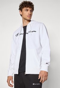 Hvit zip-up hettegenser laget av mykt stoff, med en svart «Champion» logo på forsiden, kengurulommer og snor i hetten.