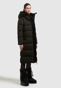 Lang sort pufferjakke med hætte, der har et quiltet design, lynlås og sidelommer. Sammen med sorte snørebånd vinterstøvler.