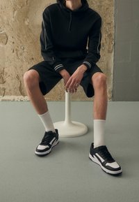 Felpa nera con strisce bianche, pantaloni corti neri, calzini bianchi e scarpe da ginnastica nere con accenti bianchi. Superficie liscia e sfondo uniforme.