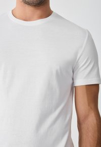 Camiseta blanca hecha de algodón suave, con un cuello redondo clásico y mangas cortas. Textura suave; sin patrones ni acentos visibles.