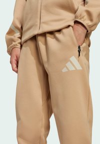 Bež flis trenirke s elastičnom pasicom, bočnim džepovima sa zipom i bijelim Adidas logotipom. Glatka tekstura s opuštenim stilom.