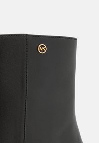 Czarny skórzany botek za kostkę o gładkiej fakturze, z widocznym złotym okrągłym logo po boku, widocznymi szwami i eleganckim designem.