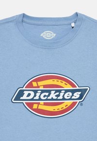 Ανοιχτόμπλε βαμβακερό t-shirt με κυκλικό λογότυπο Dickies σε κόκκινο και κίτρινο, με μαύρο και λευκό κείμενο, στρογγυλή λαιμόκοψη και κοντά μανίκια.
