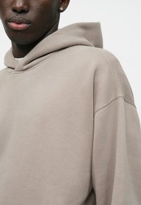 Μπεζ φούτερ με μεγάλη, μαλακή κουκούλα από ύφασμα, χαμηλωμένα ώμους και απαλό texture. Διαθέτει απλό σχέδιο και διακριτές ραφές.