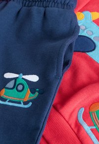 Pantalon bleu pour enfants avec un écusson d’hélicoptère vert et orange sur un tissu rouge à motif bleu et jaune.