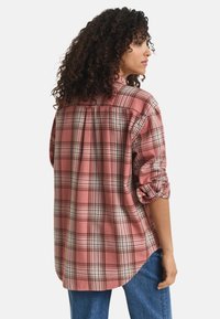 Vrouw met krullend haar, gekleed in een roze geruit shirt met lange mouwen en blauwe spijkerbroeken, iets naar de zijkant gedraaid met opgerolde mouwen.