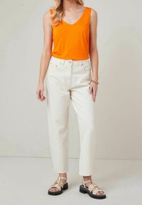 Top arancione senza maniche abbinato a jeans bianchi a vita alta. Sandalo strutturato con cinturini e suola nera spessa completa l'outfit.