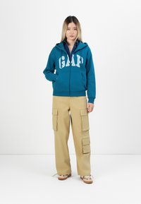 Teal zip-up hoodie with white "GAP" lettering, paired with khaki cargo pants featuring side pockets and a relaxed fit.