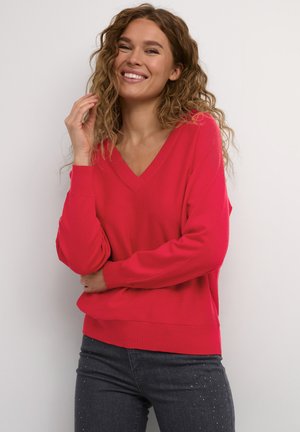 Maglione rosso a scollo a V con polsini e orlo a costine, caratterizzato da una vestibilità comoda. Indossato con jeans scuri decorati con piccoli dettagli.