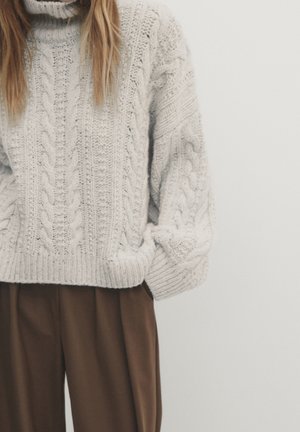 Personne portant un pull épais en maille torsadée gris clair et un pantalon ample marron, debout les mains dans les poches devant un fond uni.