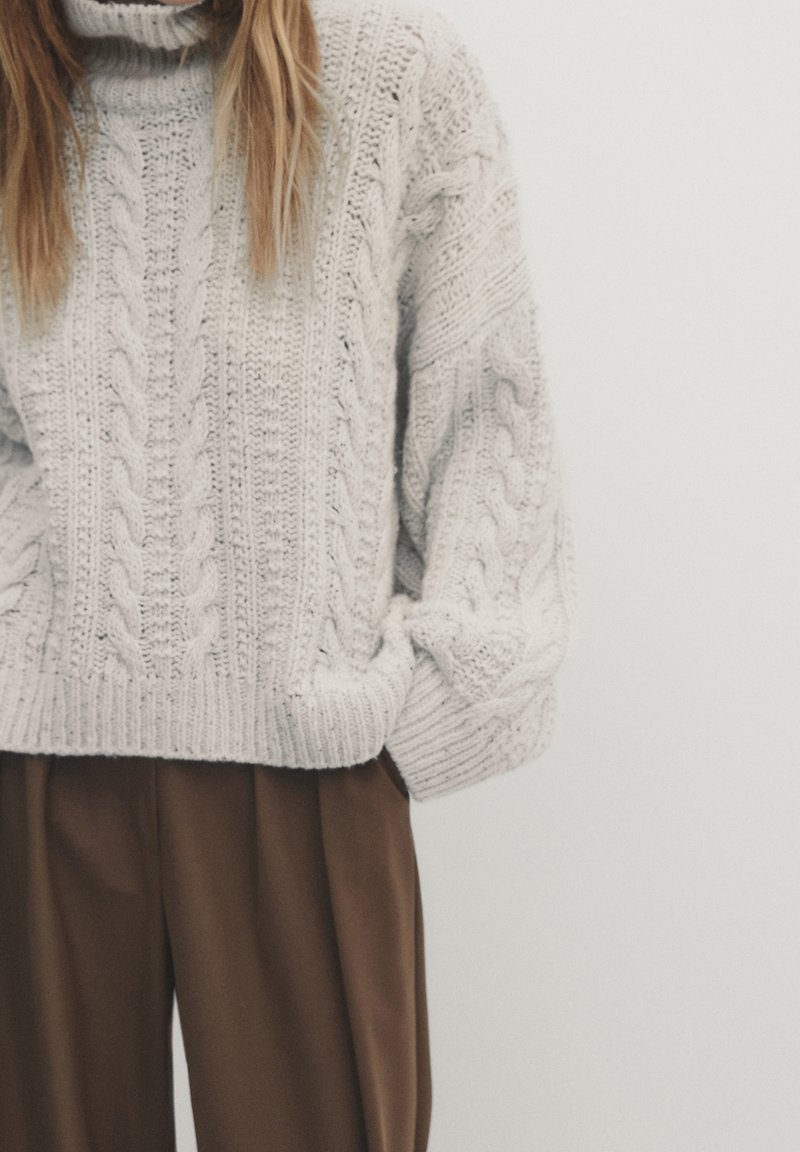 Personne portant un pull épais en maille torsadée gris clair et un pantalon ample marron, debout les mains dans les poches devant un fond uni.