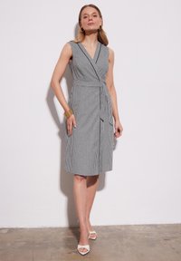 Czarna i biała sukienka w kratę typu wrap z dekoltem w serek, bez rękawów, z przednimi kieszeniami, wiązanym w pasie i długości do kolan. Noszona z białymi butami.