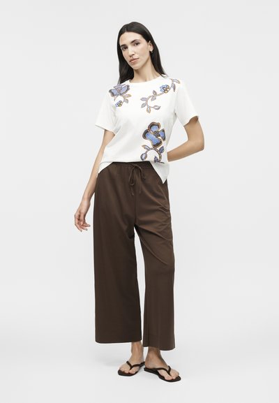 Γυναίκα που φοράει λευκό κοντομάνικο πουκάμισο με μπλε floral σχέδια, καφέ παντελόνι φαρδιάς γραμμής με κορδόνι και μαύρα σαγιονάρες, στέκεται μπροστά σε λευκό φόντο.