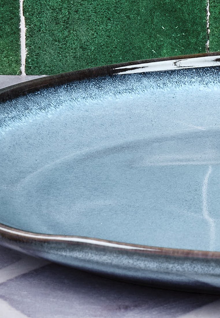 Piatto in ceramica con superficie lucida di un blu chiaro, sfumatura sottile e bordo scuro. Sullo sfondo è presente un muro piastrellato verde e bianco.