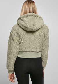 Korte olijfgroene hoodie gemaakt van zacht, textuur materiaal. Heeft een grote capuchon en ribgebreide manchetten. Draagt met zwarte aansluitende leggings.