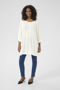 Weißes, plissiertes Tunika-Top mit rundem Ausschnitt, Dreiviertelärmeln und einer Knopfleiste vorne, kombiniert mit blauen Skinny-Jeans und schwarzen Stilettos.
