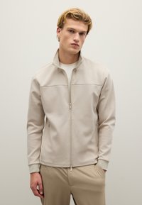 Chaqueta crema con cremallera hecha de una tela suave, con dos bolsillos laterales y un cuello alzado. Textura suave, diseño ajustado y costuras minimalistas.